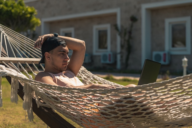 Joven con gorra tumbado en una hamaca con laptop