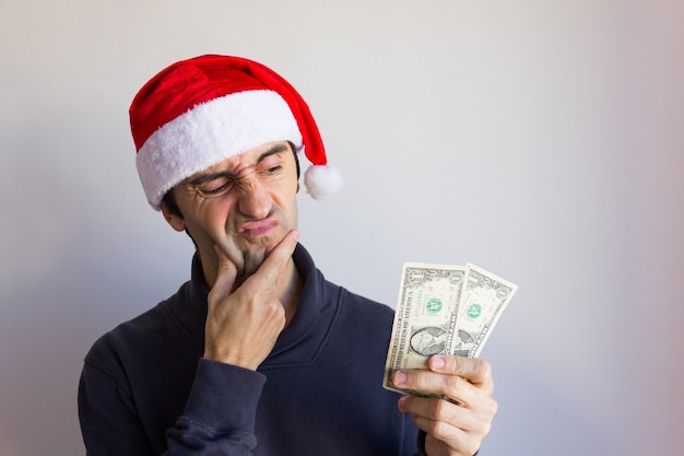 Joven con gorro de Papá Noel sosteniendo billetes de un dólar con mirada de disgusto en Navidad seaso