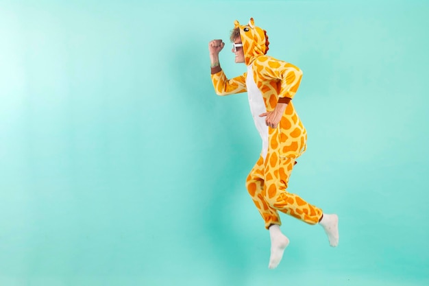 Un joven gracioso ganador con pijama de jirafa naranja salta a la fiesta con gafas de sol de fondo azul