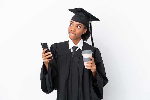 Joven graduada universitaria afroamericana aislada sobre fondo blanco sosteniendo café para llevar y un móvil mientras piensa en algo