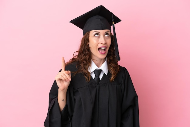 Joven graduada universitaria aislada de fondo rosa apuntando hacia arriba y sorprendida