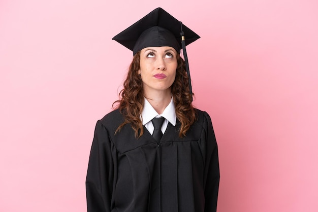 Joven graduada universitaria aislada de fondo rosa y mirando hacia arriba