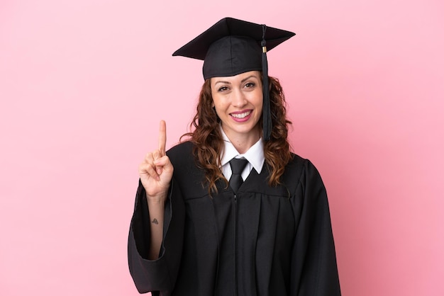 Joven graduada universitaria aislada de fondo rosa mostrando y levantando un dedo en señal de lo mejor