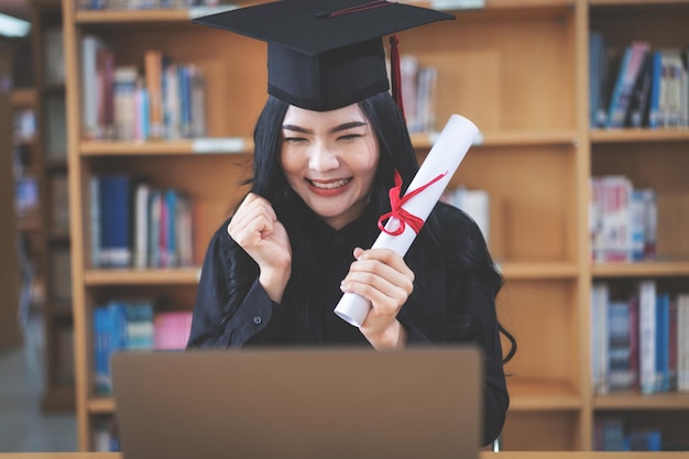 Una joven graduada universitaria asiática que expresa alegría y entusiasmo por celebrar su graduación de grado frente a una computadora portátil haciendo una videollamada remota a sus padres en casa
