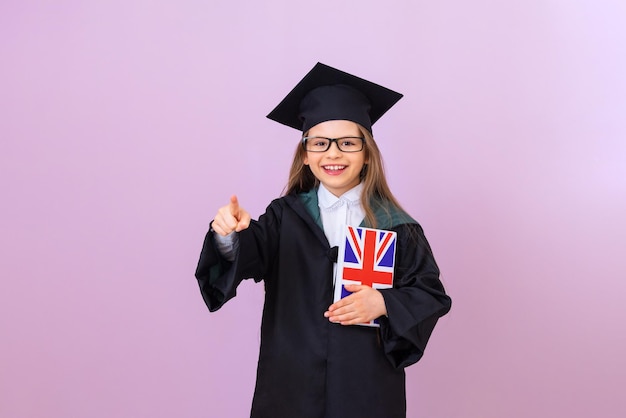 Un joven graduado con un impermeable ceremonial y un sombrero de graduación cuadrado sostiene un libro de inglés y señala hacia adelante. una colegiala sobre un fondo aislado.