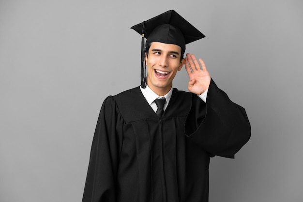 Joven graduado universitario argentino aislado de fondo gris escuchando algo poniendo la mano en la oreja