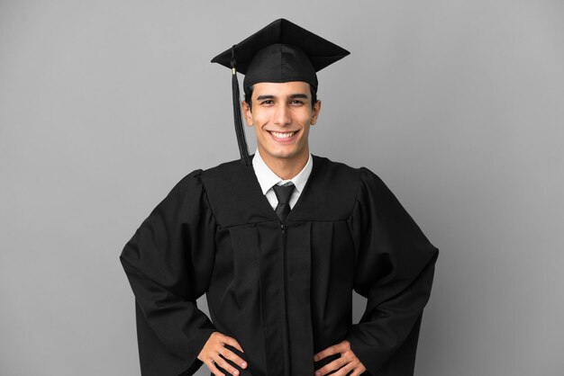 Foto joven graduado universitario argentino aislado de fondo gris posando con los brazos en la cadera y sonriendo