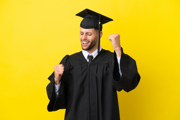 Joven graduado universitario hombre caucásico aislado sobre fondo amarillo celebrando una victoria
