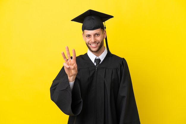 Joven graduado universitario hombre caucásico aislado sobre fondo amarillo feliz y contando tres con los dedos