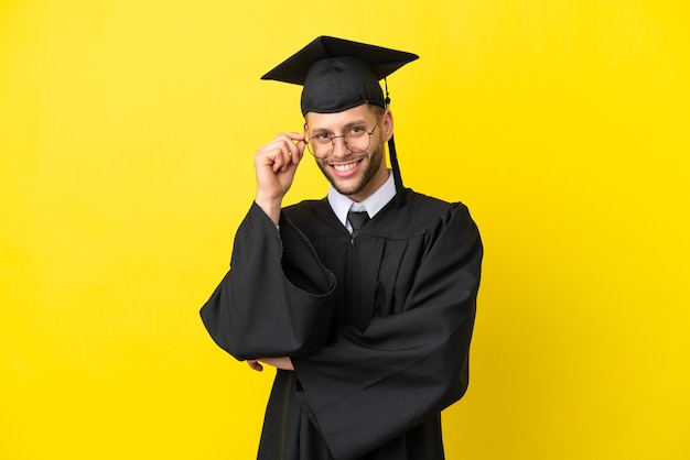 Joven graduado universitario hombre caucásico aislado sobre fondo amarillo con gafas y feliz