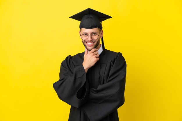 Joven graduado universitario hombre caucásico aislado sobre fondo amarillo con gafas y sonriendo