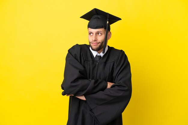 Joven graduado universitario hombre caucásico aislado sobre fondo amarillo haciendo gesto de dudas mientras levanta los hombros