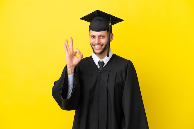 Joven graduado universitario hombre caucásico aislado sobre fondo amarillo mostrando un signo de ok con los dedos