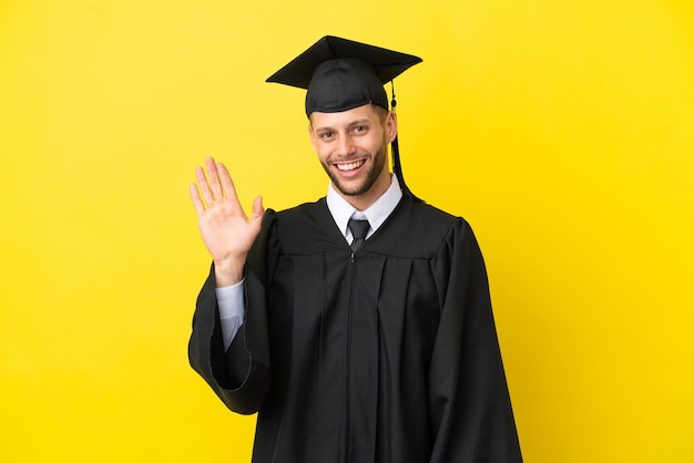 Joven graduado universitario hombre caucásico aislado sobre fondo amarillo saludando con la mano con expresión feliz