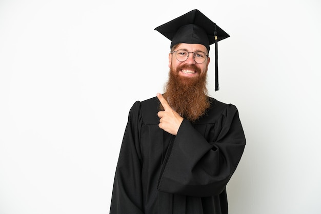 Joven graduado universitario hombre rojizo aislado sobre fondo blanco apuntando hacia un lado para presentar un producto