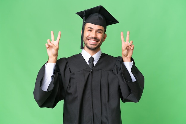 Joven graduado universitario sobre un fondo aislado que muestra el signo de la victoria con ambas manos