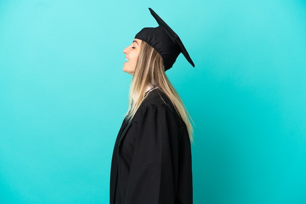 Joven graduado universitario sobre fondo azul aislado riendo en posición lateral