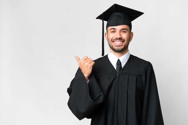 Joven graduado universitario sobre fondo blanco aislado apuntando hacia un lado para presentar un producto