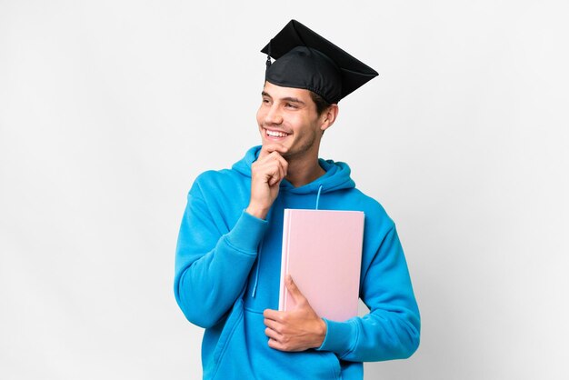 Joven graduado universitario sobre fondo blanco aislado mirando hacia arriba mientras sonríe