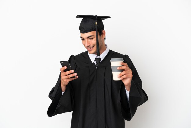 Joven graduado universitario sobre fondo blanco aislado sosteniendo café para llevar y un móvil