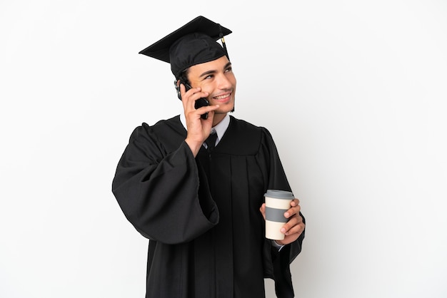 Joven graduado universitario sobre fondo blanco aislado sosteniendo café para llevar y un móvil