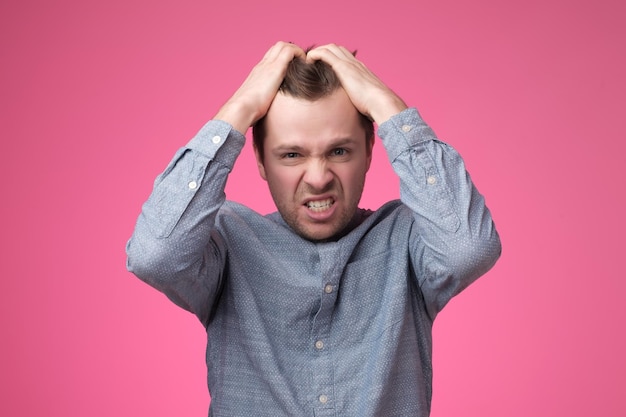 Foto joven gritando enojado poniendo las manos en la cabeza en la pared rosa en el estudio