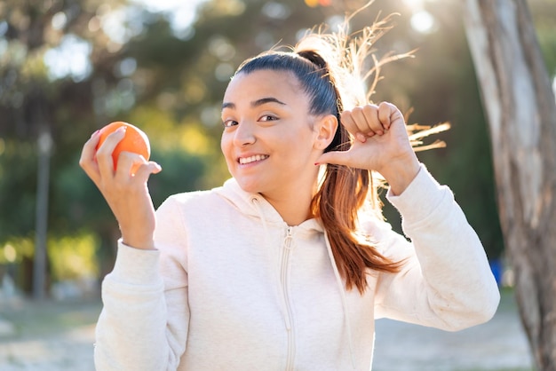 Joven guapa morena sosteniendo una naranja al aire libre orgullosa y satisfecha