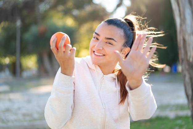 Joven guapa morena sosteniendo una naranja al aire libre saludando con la mano con expresión feliz