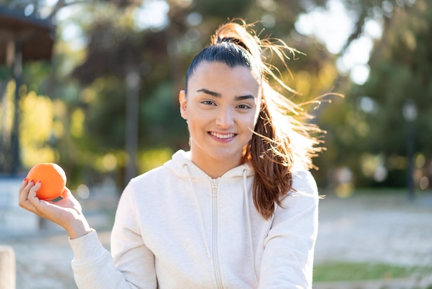 Joven guapa morena sosteniendo una naranja al aire libre sonriendo mucho