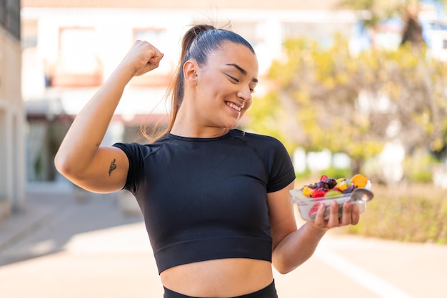 Foto joven guapa morena sosteniendo un plato de fruta al aire libre celebrando una victoria