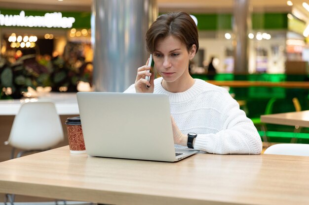 Joven guapa morena con sudadera blanca sentada en un centro comercial en una mesa y trabajando en una computadora portátil, usando un teléfono móvil. Concepto independiente y de negocios.