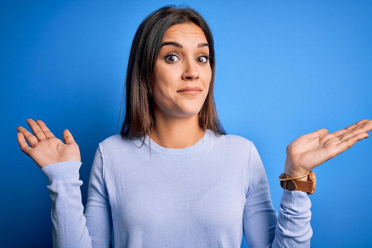 Foto joven guapa morena con suéter casual sobre fondo azul expresión despistada y confusa con los brazos y las manos levantadas concepto de duda