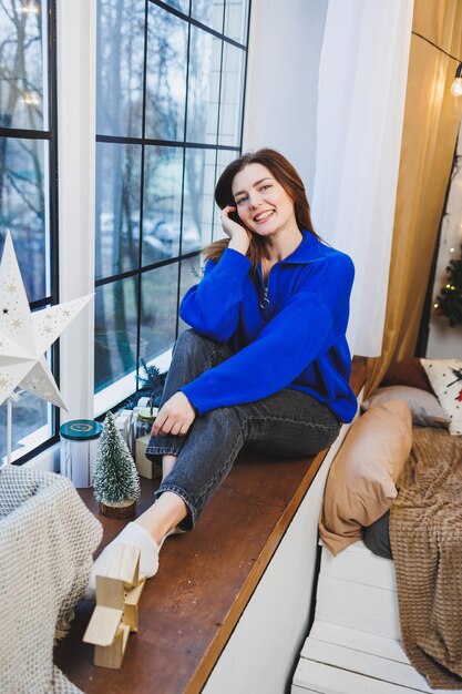 Una joven guapa con un suéter de punto azul está sentada junto a la ventana esperando el nuevo año el estado de ánimo de Año Nuevo en un ambiente hogareño