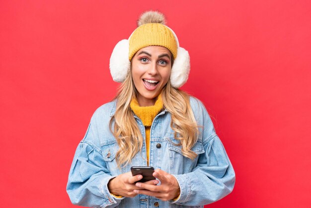 Joven guapa uruguaya con orejeras de invierno aisladas en un fondo rojo sorprendida y enviando un mensaje