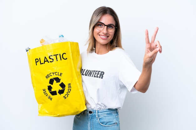 Joven guapa uruguaya sosteniendo una bolsa llena de botellas de plástico para reciclar aisladas de fondo blanco sonriendo y mostrando el signo de la victoria