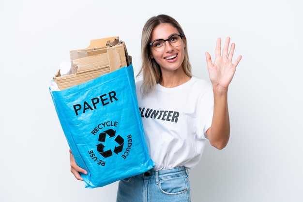 Joven guapa uruguaya sosteniendo una bolsa de reciclaje llena de papel para reciclar aislada en un fondo de croma blanco saludando con la mano con expresión feliz