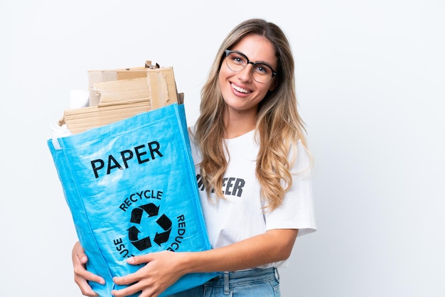 Joven guapa uruguaya sosteniendo una bolsa de reciclaje llena de papel para reciclar aislada en un fondo de croma blanco sonriendo mucho