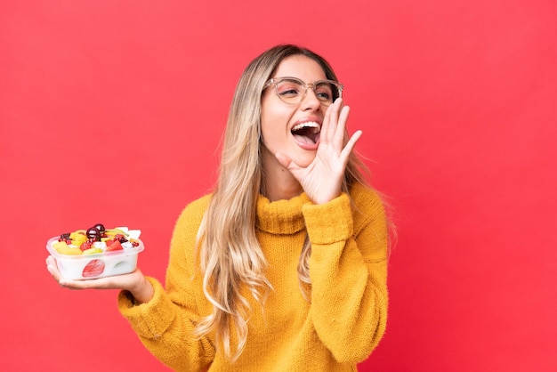 Joven guapa uruguaya sosteniendo un tazón de fruta aislada de fondo rojo gritando con la boca abierta a un lado