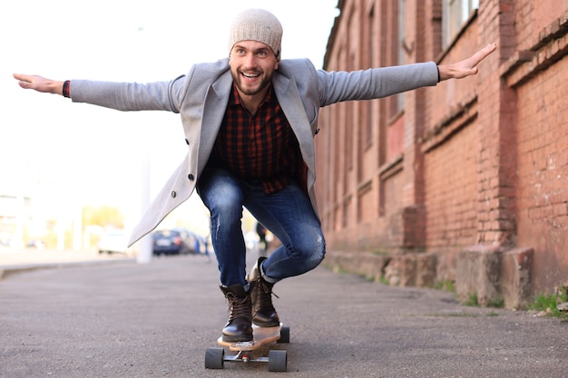 Joven guapo con abrigo gris y sombrero en longboard en la calle de la ciudad. Concepto de skate urbano.