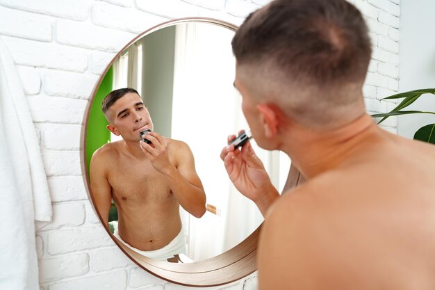 Un joven guapo se afeita la cara con una afeitadora eléctrica sobre un fondo blanco