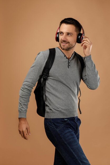 Un joven guapo con auriculares y mochila escucha música aislado en un fondo beige