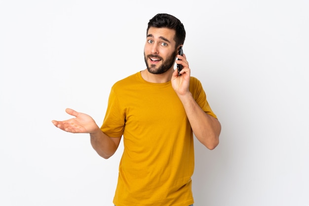 Joven guapo con barba aislado en la pared blanca manteniendo una conversación con el teléfono móvil con alguien