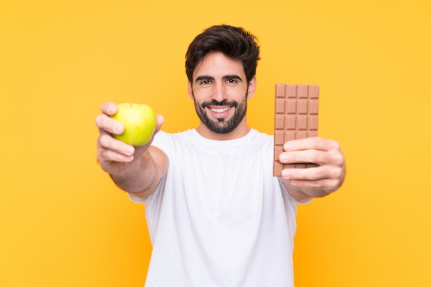 Joven guapo con barba sobre pared amarilla aislada tomando una tableta de chocolate en una mano y una manzana en la otra