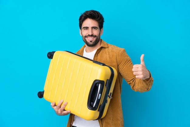 Joven guapo con barba sobre pared azul aislada en vacaciones con maleta de viaje y con el pulgar hacia arriba