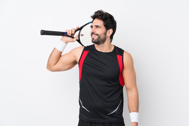 Joven guapo con barba sobre pared blanca aislada jugando tenis y mirando hacia arriba