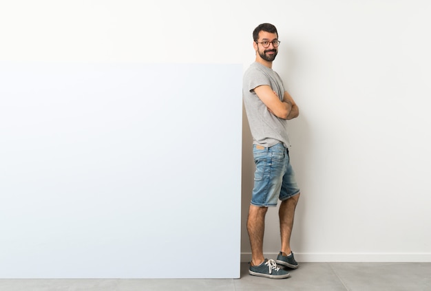 Joven guapo con barba sosteniendo un gran cartel azul vacío con gafas y sonriendo