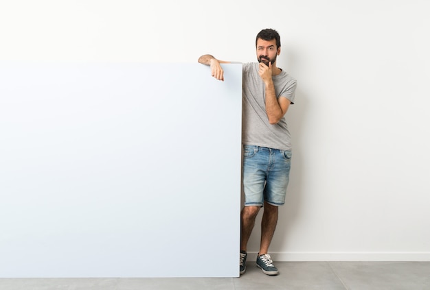 Joven guapo con barba sosteniendo un gran cartel vacío azul pensando