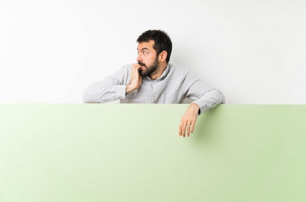 Joven guapo con barba sosteniendo un gran cartel vacío nervioso y asustado