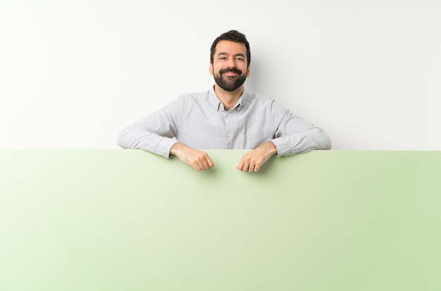 Joven guapo con barba sosteniendo un gran cartel verde vacío posando con los brazos en la cadera y sonriendo