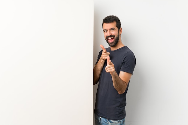 Joven guapo con barba sosteniendo una gran pancarta vacía apuntando hacia el frente y sonriendo